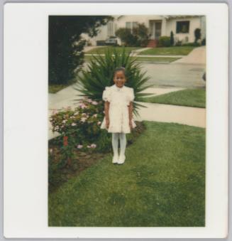 [Young girl in white dress, white stockings and Mary-Jane shoes standing outside on lawn in front of tropical plant]