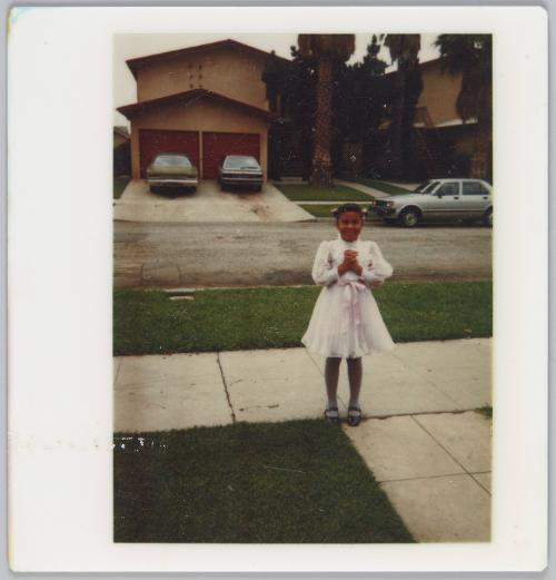 [Girl in light pink dress with ribbon tied around her waist standing on sidewalk next to grass lawn]