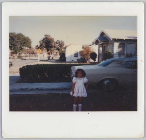 [Young girl wearing pink and white dress and high socks standing in driveway with car behind her]

