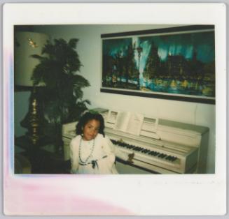 [Girl sitting at white piano wearing a white dress and blue necklace]
