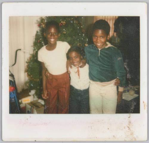 [Two boys and a girl standing with arms around each other in front of Christmas tree]
