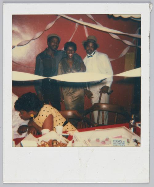 [Two men and a woman standing together and smiling at party, streamers hung on walls]
