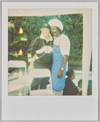 [Man in green and white tracksuit and "M" baseball cap with arm around man in white hat and blue apron, outside by white folding chairs]