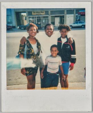[Man with his arm around a woman to the left and a teenage boy to the right, boy in striped t-shirt standing in front, they are on the sidewalk]