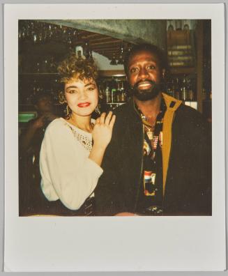 [Woman standing beside man at a bar, she has her hand on his shoulder]