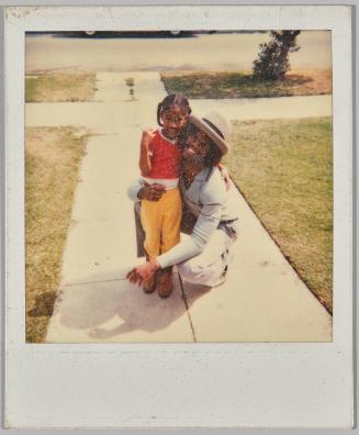 [Girl in yellow pants being hugged by man in white brimmed hat crouched beside her, they are outside]