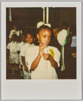 [Girl in white dress and white hair ties with a gold ribbon, other children lined up behind her]