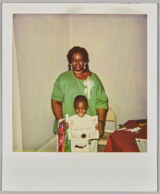 [Woman in green dress standing behind a child displaying a diploma]