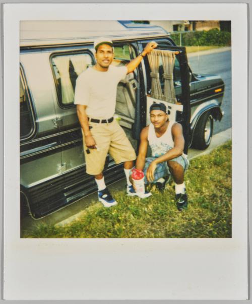 [Two men, one standing and one crouching, beside a vehicle with open passenger door]