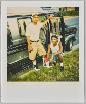 [Two men, one standing and one crouching, beside a vehicle with open passenger door]
