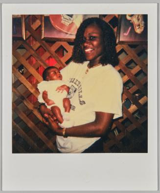 [Woman in white t-shirt holding baby, standing inside in front of wood lattice wall with framed pictures]