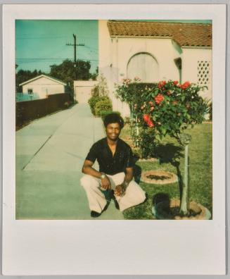 [Man in black shirt and white pants crouching in driveway beside flower bushes]