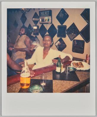 [Woman with her hair pulled back sitting behind a bar, wall behind her is decorated with reflective squares and diamonds]