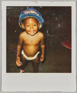 [Toddler in diaper and oversized blue baseball cap, smiling at camera]