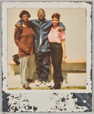 [Man with arms around two women standing in front of wooden bench and white wall]