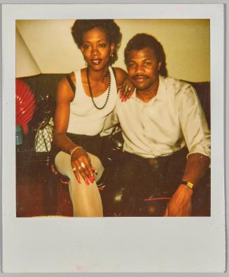 [Man and woman sitting next to each other, woman is wearing a beaded necklace and has red fingertips]