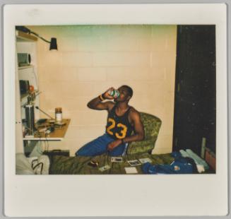 [Man sitting at desk with Polaroids on bed, drinking out of can]
