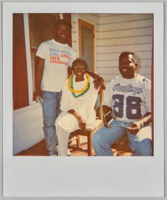 [Two men and elderly woman sitting on porch, man on right wearing a graphic shirt "Rawlings 88"]
