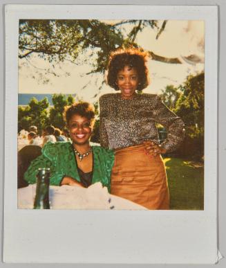 [Woman in green blazer and necklace sitting at a table outside, she has her arm around the waist of a woman standing beside her]