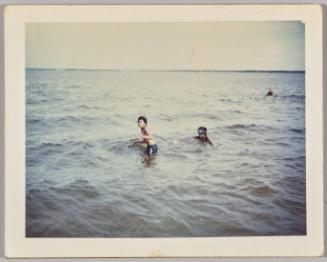 [Two children wading in open body of water, child on right is wearing goggles]
