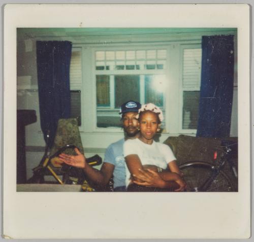 [Man sitting with woman wearing curlers in her hair on his lap with large window in background]
