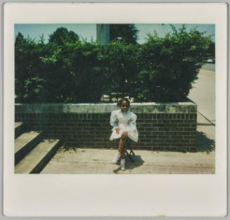 [Little girl in white dress sitting on stool outside in concrete area next to stairwell]

