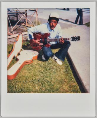 [Man sitting on grass with guitar in hands and guitar case on the left side]
