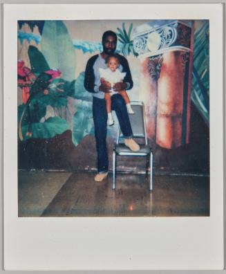 [Man standing with one leg raised onto seat of chair with baby balanced on his knee]
