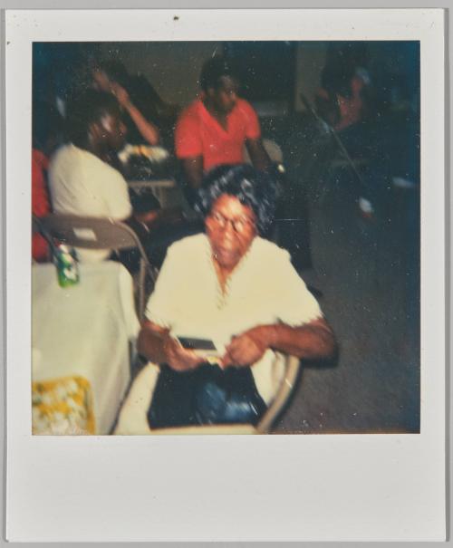 [Blurry photograph of elderly woman wearing glasses and white short sleeve top]
