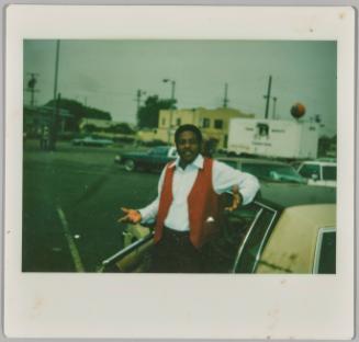 [Man standing in doorway of car with arms slightly raised in shrugging gesture]
