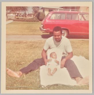[Man sitting on white towel on lawn with baby between his legs sleeping in car seat]
