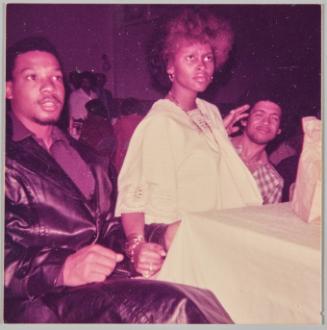 [Man and woman sitting at table, man in the background squinting his eyes]
