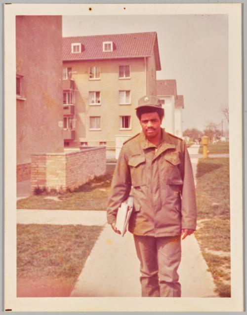 [Man in green uniform holding binders at his side walking down sidewalk]
