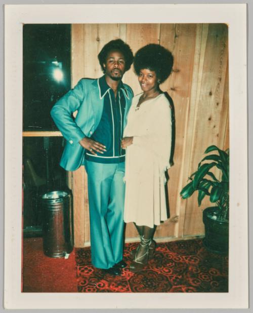 [Man in green suit posing with woman in white in wood panelled room with red carpet]

