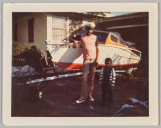Leonard & Tyrone, Oct 25, 73 [Man and little boy posing in front of boat]