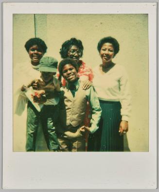 [Three women with two boys posing in formal outfits against concrete wall]
