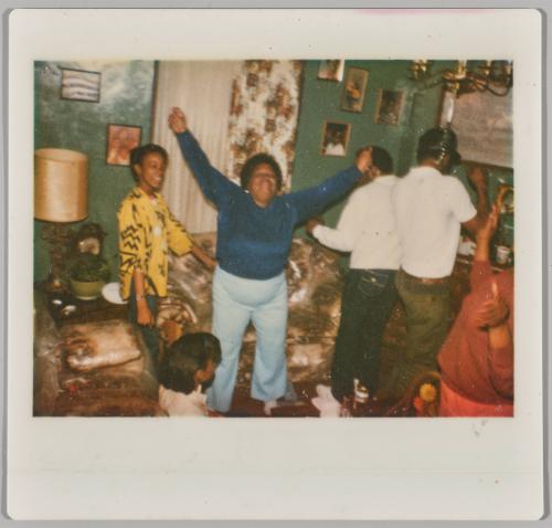 [Group of people dancing in living room, woman in centre has arms raised in air]
