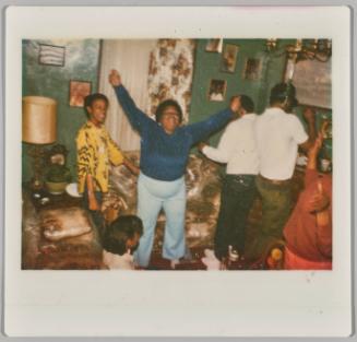 [Group of people dancing in living room, woman in centre has arms raised in air]
