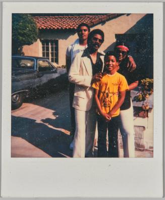 [Man, woman, adolescent boy and young boy posing in front of house and car]
