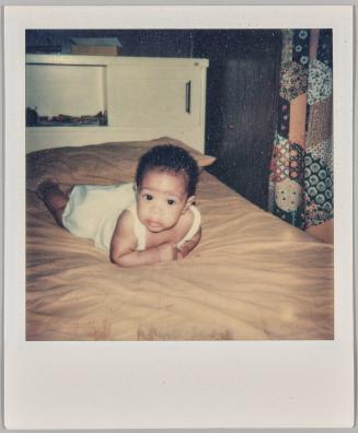 [Baby in white dress laying on stomach on beige bed]
