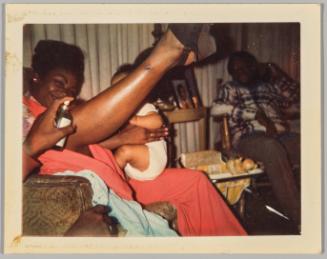 [Woman seated with legs elevated holding baby, hand entering frame with perfume bottle]
