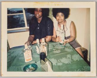 [Two women at table with bottles of gin, one woman holding bottle]

