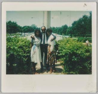 [Man and two women dressed formally standing in front of parking lot]
