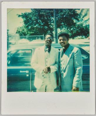 [Two men dressed formally standing beside car, man on left is a priest]
