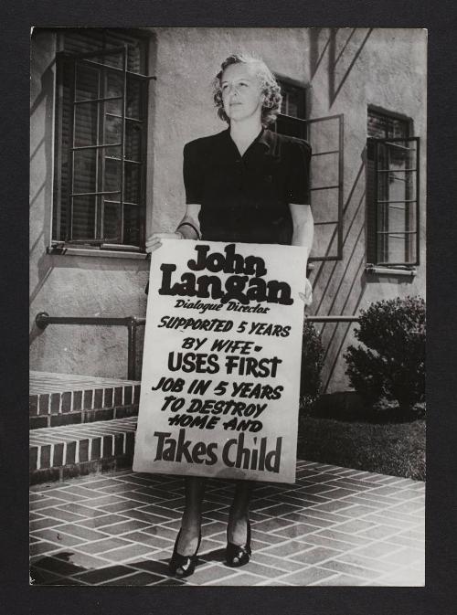 Woman holding up a sign - "John Langan, dialogue director, supported 5 years by wife…"