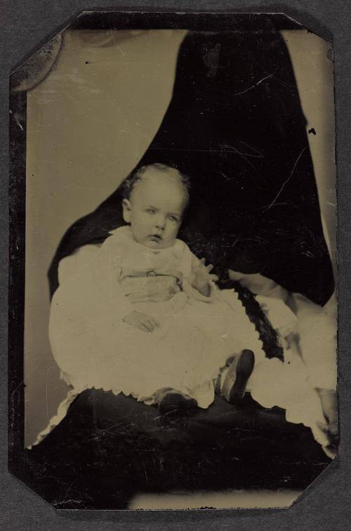 Portrait of baby in long white dress (seated on lap of shrouded figure)