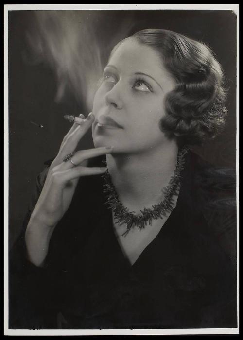 Woman smoking, looking up, wearing a black top and necklace.