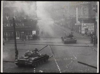 Driving into action in Budapest. At the corner people stand watching. Smoke rises in the background.