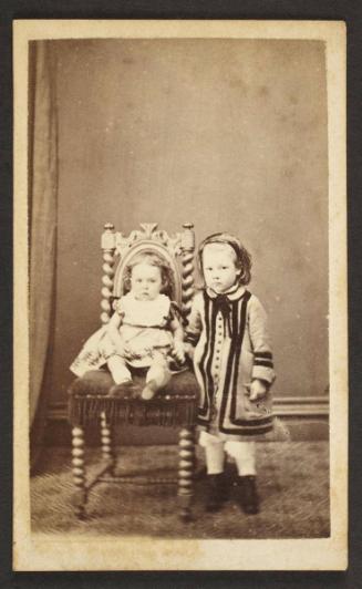Unknown sitter [portrait of seated infant on upholstered chair left to a standing girl]
