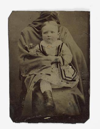 Portrait of a young girl wearing boots (seated on lap of shrouded figure)